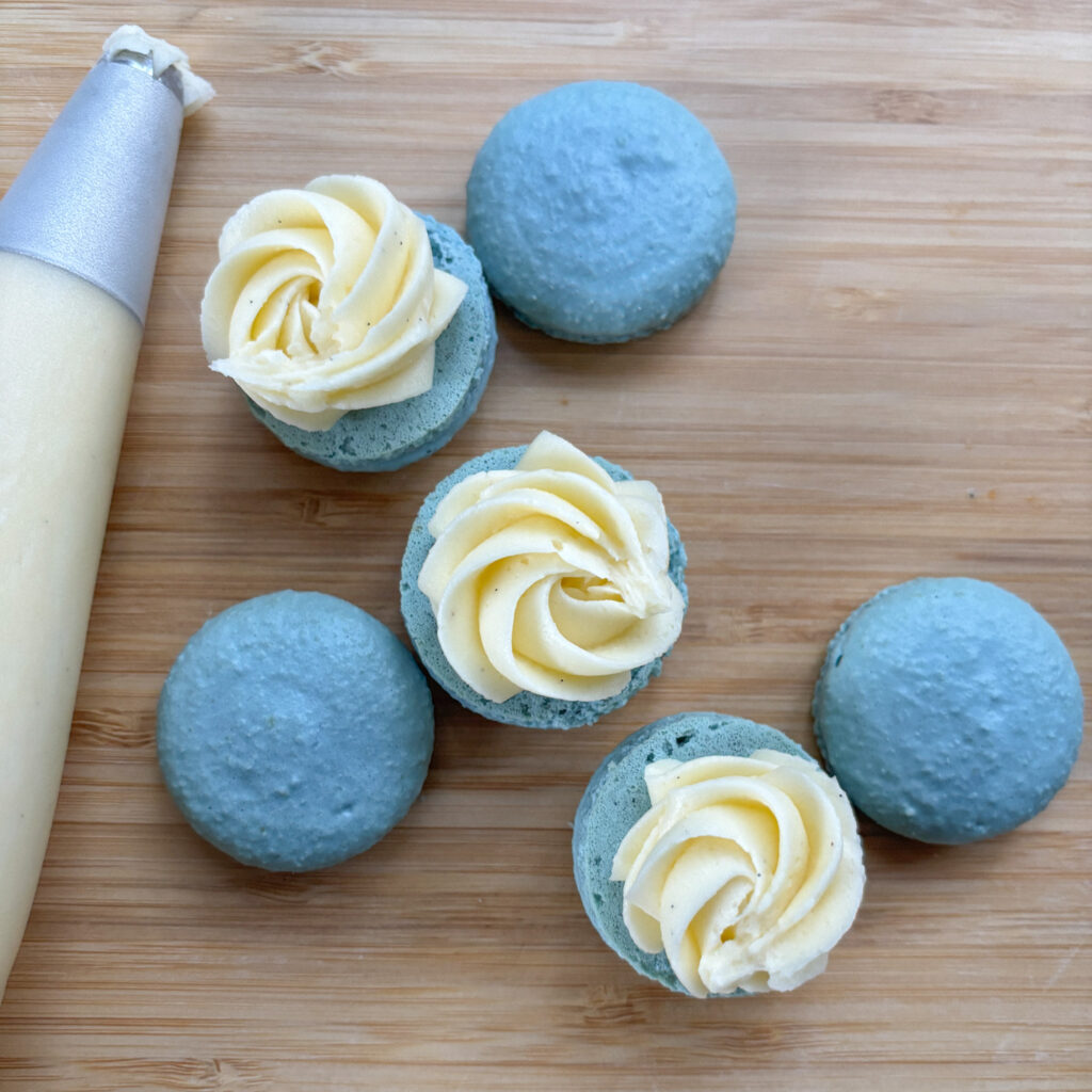 Vanilla Buttercream for Macarons showing buttercream in piping bag and blue macarons being filled with star tip